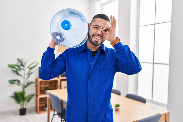 Homem de serviço hispânico segurando uma garrafa de galão de água para entrega sorrindo feliz fazendo sinal ok com a mão no olho olhando através dos dedos  - Foto, Imagem