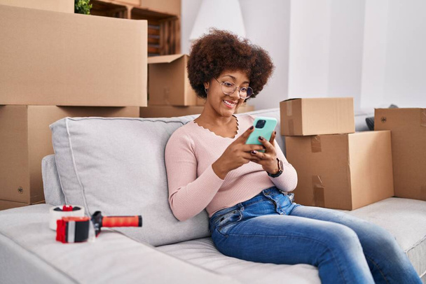 Femme afro-américaine utilisant un smartphone assis sur le canapé à la maison - Photo, image