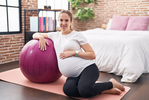 Νεαρή έγκυος γυναίκα αγγίζει την κοιλιά ακουμπά σε fit μπάλα στο υπνοδωμάτιο - Φωτογραφία, εικόνα