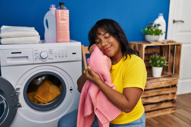 Young beautiful latin woman washing clothes touching face with soft sweater at laundry room - Fotoğraf, Görsel