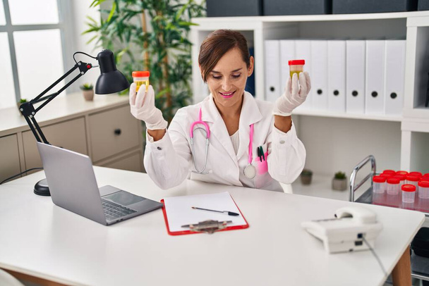 Middle age woman wearing doctor uniform holding urine analysis test tubes at clinic - Photo, Image