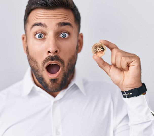 Knappe Spaanse man met cardano cryptogeld munt bang en verbaasd met open mond voor verrassing, ongeloof gezicht  - Foto, afbeelding