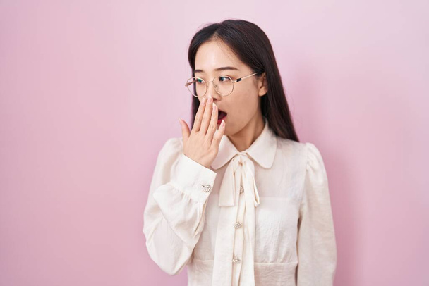 Молодая китаянка, стоящая на розовом фоне, устала зевать, прикрывая рот рукой. беспокойство и сонливость.  - Фото, изображение