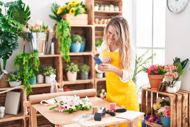 Young blonde woman florist make photo to flowers by smartphone at florist - 写真・画像