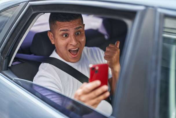 Jeune homme hispanique faisant appel vidéo avec smartphone dans la voiture pointant le pouce vers le côté souriant heureux avec la bouche ouverte  - Photo, image