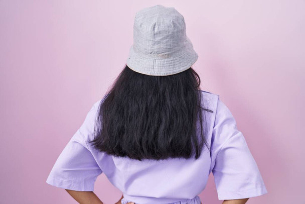 Young hispanic woman standing over pink background wearing hat standing backwards looking away with arms on body  - Fotó, kép