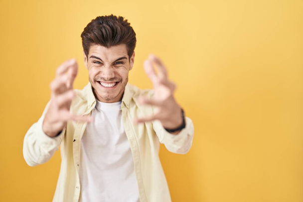 Jonge Spaanse man die over een gele achtergrond staat te schreeuwen gefrustreerd door woede, handen die proberen te wurgen, gek schreeuwend  - Foto, afbeelding