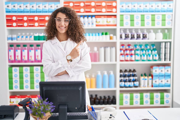 Mulher hispânica com cabelo encaracolado trabalhando na farmácia farmácia com um grande sorriso no rosto, apontando com o dedo da mão para o lado olhando para a câmera.  - Foto, Imagem