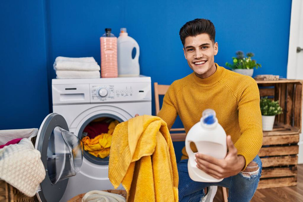 Young hispanic man washing clothes holding detergent bottle at laundry - Foto, imagen