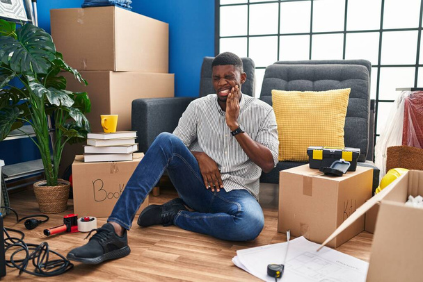 Yeni evinde yerde oturan Afrikalı Amerikalı bir adam diş ağrısı ya da diş ağrısı yüzünden acı dolu bir ifadeyle ağzına dokunuyor. dişçi  - Fotoğraf, Görsel