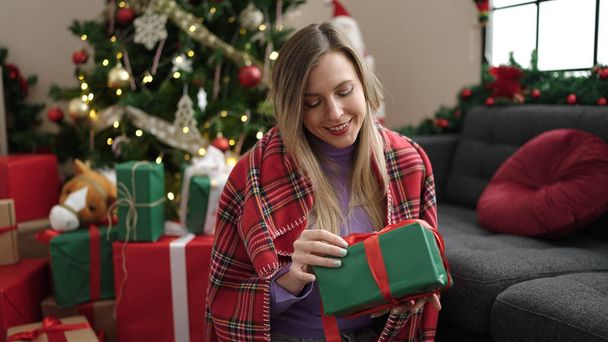 Νεαρή ξανθιά γυναίκα κρατώντας δώρο κάθεται στο πάτωμα από το χριστουγεννιάτικο δέντρο στο σπίτι - Φωτογραφία, εικόνα