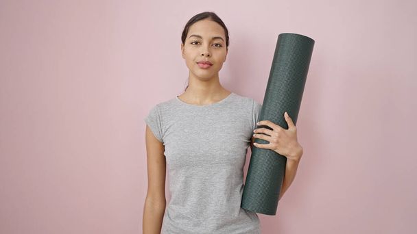 Jonge mooie latino vrouw dragen sportkleding houden yoga mat over geïsoleerde roze achtergrond - Foto, afbeelding