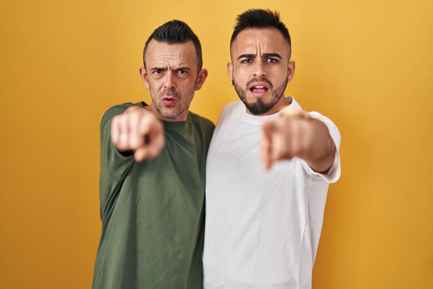 Homosexuální pár stojící nad žlutým pozadím ukazuje nespokojenost a frustraci na kameru, naštvaný a zběsilý na vás  - Fotografie, Obrázek