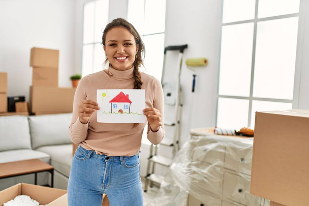 Joven hermosa mujer hispana sonriendo confiado sosteniendo casa dibujar en nuevo hogar - Foto, imagen