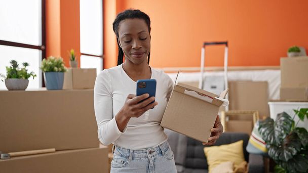 Afričanky americká žena pomocí smartphone držení balíček v novém domově - Fotografie, Obrázek