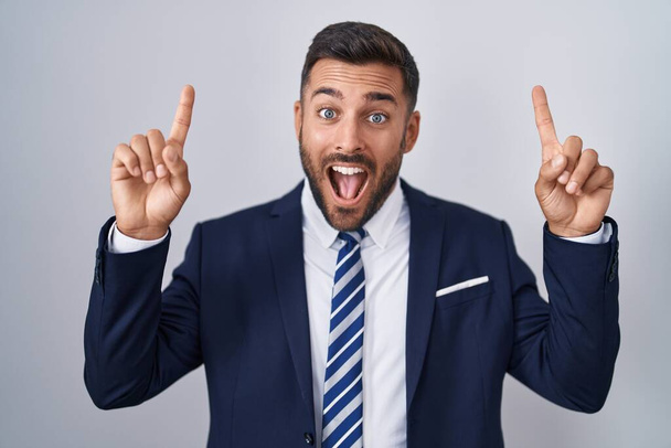 Hombre hispano guapo vistiendo traje y corbata sonriendo asombrado y sorprendido y señalando con los dedos y los brazos levantados.  - Foto, Imagen
