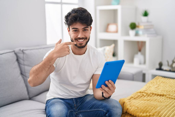 Homem hispânico com barba usando touchpad sentado no sofá sorrindo alegre mostrando e apontando com os dedos dentes e boca. conceito de saúde dentária.  - Foto, Imagem