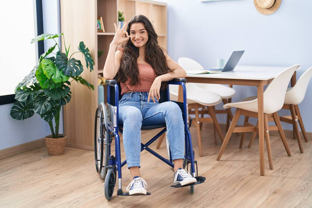 Молодая девушка-подросток, сидящая на инвалидной коляске в гостиной делает знак ОК с пальцами, улыбаясь дружелюбный жест отличный символ  - Фото, изображение