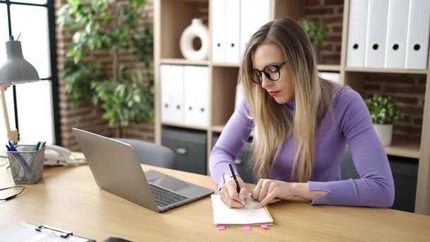 Junge blonde Geschäftsfrau schreibt im Büro mit Laptop auf Notizbuch - Foto, Bild