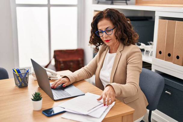 Middelbare leeftijd Spaanse vrouw werken met laptop en documenten op kantoor - Foto, afbeelding
