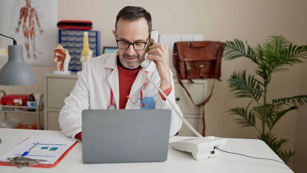 Középkorú férfi orvos használja laptop beszél telefonon a klinikán - Fotó, kép