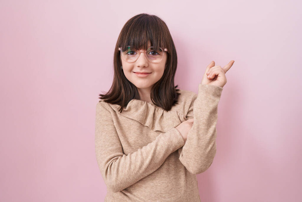 Μικρό ισπανόφωνο κορίτσι που φοράει γυαλιά χαμογελώντας χαρούμενο δείχνοντας με το χέρι και το δάχτυλο στο πλάι  - Φωτογραφία, εικόνα