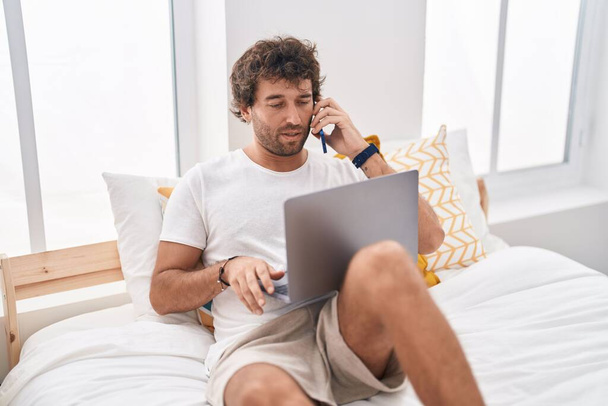 Young hispanic man using laptop talking on smartphone at bedroom - Foto, immagini