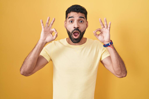 Hombre hispano con barba de pie sobre fondo amarillo mirando sorprendido y sorprendido haciendo símbolo de aprobación bien con los dedos. expresión loca  - Foto, imagen