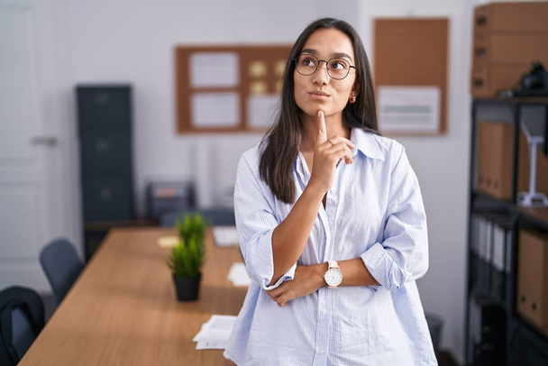 Junge hispanische Frau im Büro, die konzentriert mit dem Finger am Kinn über Zweifel nachdenkt und fragend nach oben schaut  - Foto, Bild