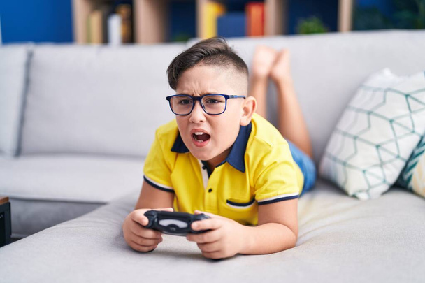 Jeune enfant hispanique jouant contrôleur de tenue de jeu vidéo sur le canapé en colère et fou hurlant frustré et furieux, criant de colère. rage et concept agressif.  - Photo, image