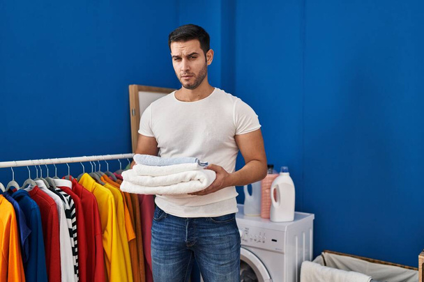 Young hispanic man with beard holding folded clean towels at laundry room skeptic and nervous, frowning upset because of problem. negative person.  - Foto, immagini