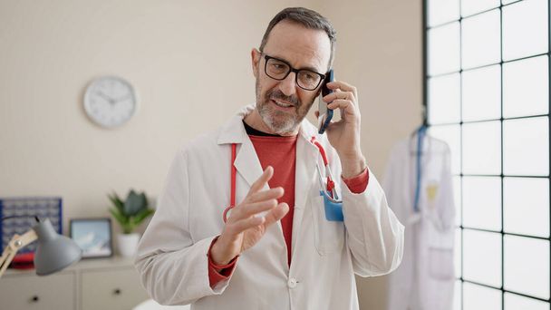 Medico uomo di mezza età sorridente fiducioso parlando su smartphone in clinica - Foto, immagini
