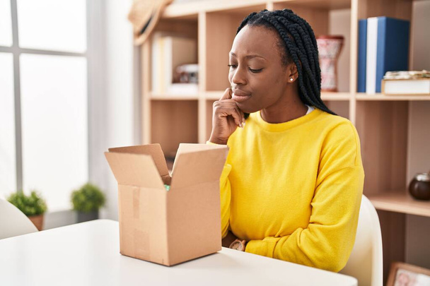 Beautiful black woman opening cardboard box thinking concentrated about doubt with finger on chin and looking up wondering  - 写真・画像