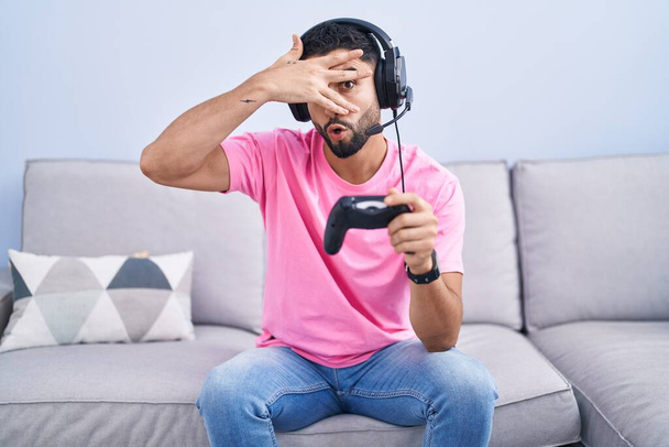 Hispanic young man playing video game holding controller sitting on the sofa peeking in shock covering face and eyes with hand, looking through fingers with embarrassed expression.  - Foto, Imagen
