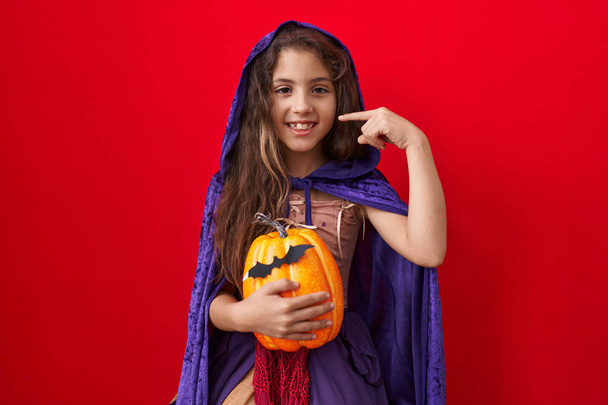 Mała Latynoska dziewczyna w kostiumie czarownicy halloween wskazującym palcem na siebie uśmiechniętą szczęśliwą i dumną  - Zdjęcie, obraz