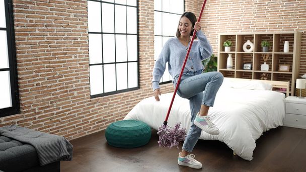 Young beautiful hispanic woman smiling confident using mop as a guitar at home - 写真・画像