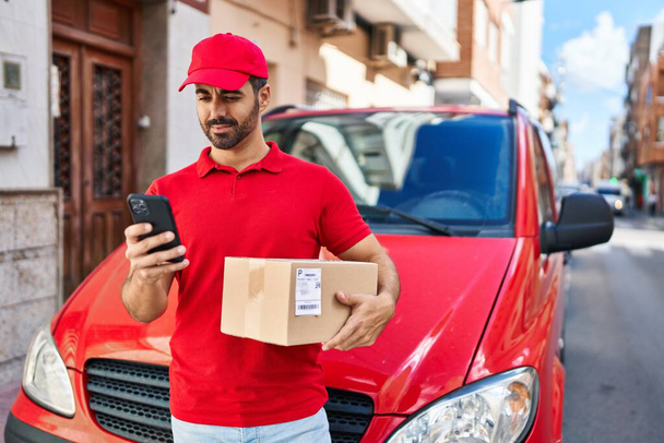 Young hispanic man courier using smartphone holding package at street - Foto, afbeelding