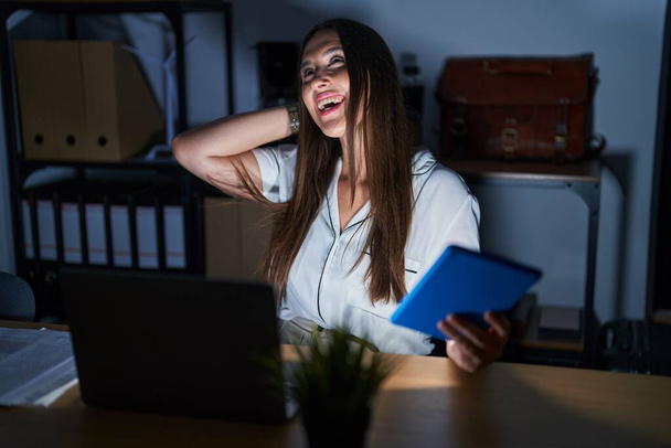 Giovane donna bruna che lavora in ufficio di notte sorridendo fiduciosa toccando i capelli con gesto mano in su, in posa attraente e alla moda  - Foto, immagini