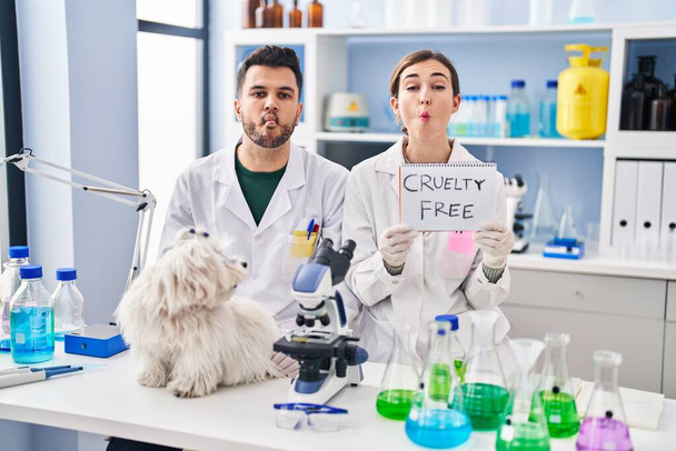 Jóvenes hispanos trabajando en laboratorio científico con perro haciendo cara de pez con boca y ojos entrecerrados, locos y cómicos.  - Foto, imagen