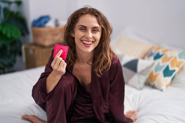 Belle femme brune tenant un préservatif dans la chambre à coucher regardant positif et heureux debout et souriant avec un sourire confiant montrant des dents  - Photo, image
