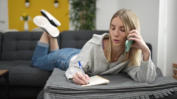 Nuori blondi nainen kirjoittaa muistikirjaan makaa sohvalla puhuu puhelimessa kotona - Valokuva, kuva