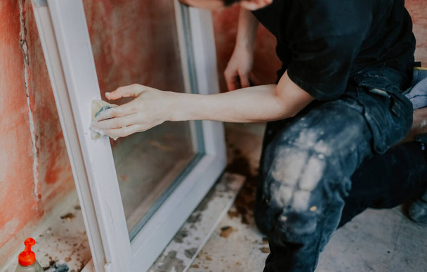 Homem jovem caucasiano lavando uma moldura da janela com uma esponja e sabão enquanto estava de pé em um joelho em um piso de azulejos velho, vista lateral close-up com foco seletivo. O conceito de renovação da casa, lavando caixilhos das janelas. - Foto, Imagem