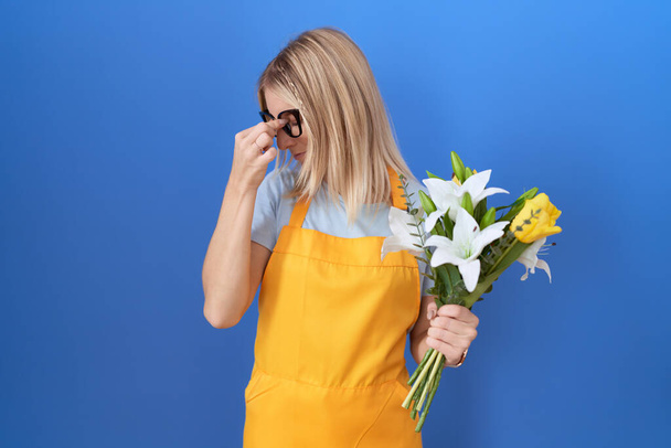 Молода біла жінка в квітковому фартусі тримає квіти втомленими, потираючи ніс і очі, відчуваючи втому і головний біль. концепція стресу та розчарування
.  - Фото, зображення