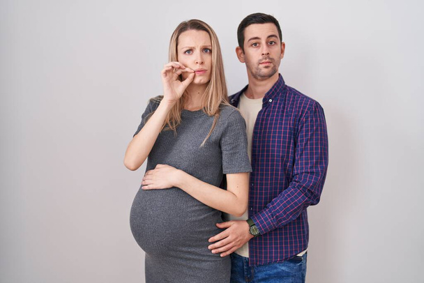 Junges Paar erwartet ein Baby, das über weißem Hintergrundmund steht und die Lippen als Reißverschluss mit Fingern geschlossen hält. Geheimniskrämerei und Schweigen, Tabuthema  - Foto, Bild