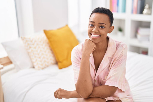 Mujer afroamericana sonriendo confiada sentada en la cama en el dormitorio - Foto, Imagen