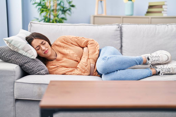 Νεαρή όμορφη ισπανόφωνη γυναίκα που υποφέρει για εμμηνορροϊκό πόνο ξαπλωμένη στον καναπέ στο σπίτι - Φωτογραφία, εικόνα