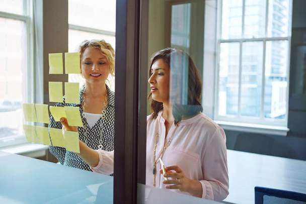 Correre attraverso le loro idee. due donne d'affari che fanno brainstorming con appunti adesivi su una parete di vetro in un ufficio - Foto, immagini