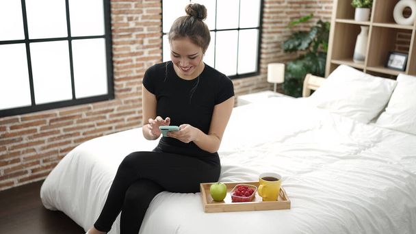 Fiatal gyönyörű spanyol nő, egészséges reggelit használ okostelefon a hálószobában - Fotó, kép