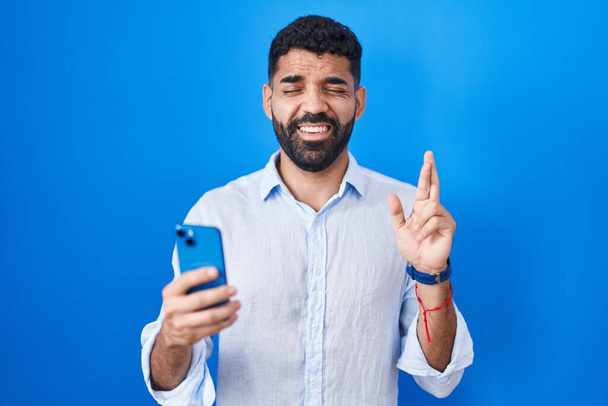 Hispanic man with beard using smartphone typing message gesturing finger crossed smiling with hope and eyes closed. luck and superstitious concept.  - Fotografie, Obrázek