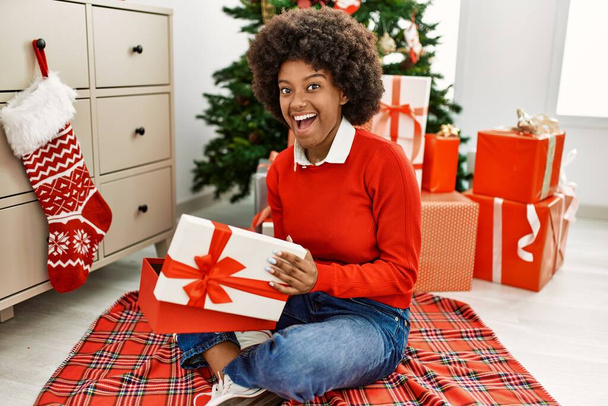 Young african american woman unboxing gift sitting by christmas tree at home - Photo, Image